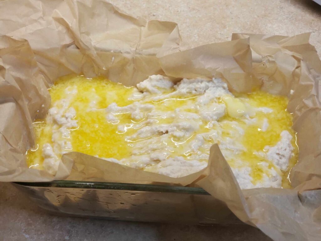 Beer Bread dough in glass loaf pan with parchment paper and butter drizzle.